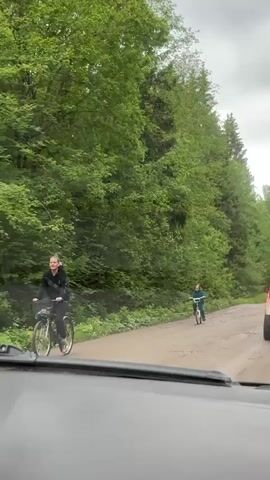From the perspective of a car driving down a dirt road, a person wearing a green jumpsuit rides a bicycle toward the car. Trees densely line the side of the road. The sky is overcast.