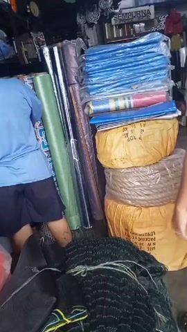 A short video clip shows an indoor scene, likely a market or storage area, filled with various goods. In the foreground, a pile of green netting is visible. Two individuals, one wearing a blue shirt and dark shorts, the other a brown t-shirt and patterned shorts, are standing near stacks of rolled materials. The rolls appear to be a mix of colors, including green, black, and blue. Further back, several large, yellow, burlap-like sacks are stacked. A man in a dark shirt and white shorts enters the frame from the right, gesturing with his hand. The environment appears somewhat cluttered, with merchandise filling the space. The lighting is dim, suggesting an indoor setting.