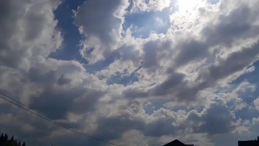 The video shows a time-lapse recording of a cloudy sky. The sun is visible through gaps in the clouds, creating a bright, hazy effect. The clouds appear to be moving and changing shape. In the lower portion of the frame, there are some dark silhouettes that could be trees or buildings, and thin wires are visible crossing the frame from left to right. The overall impression is of a dynamic sky with shifting light and cloud formations.