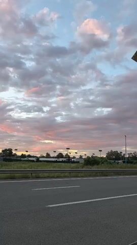The video shows a view from a car traveling on a highway. The scene is set in Saint-Aybert, France, during a sunset. The sky is filled with dramatic clouds, painted in hues of pink, purple, and blue, suggesting the end of a day. The highway stretches ahead, with a metal guardrail separating it from a grassy median. Beyond the median, a line of trees and some structures are visible, possibly parking lots or commercial buildings, illuminated by tall light poles. A striking concrete sculpture, characterized by two tall, slender pillars topped with angular, abstract shapes, dominates the right side of the frame. The sculpture is a prominent landmark in the area. The overall mood is serene and somewhat awe-inspiring, capturing the beauty of a natural phenomenon against an engineered landscape. There is no visible text in the media.