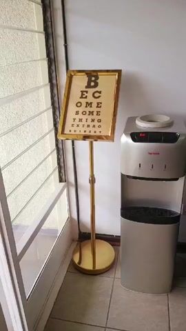 The video shows a corner of a room, focusing on a water cooler and an eye chart-style artwork. The room has a white wall on one side, against which the water cooler and the art are placed. To the left is a window with white blinds, creating vertical lines. The water cooler is a two-tone silver appliance with a black base. It has a red light on top and the brand name "Top Notch" printed on the front. Next to it is a golden-framed art piece on a stand. The art is designed like an eye chart, with the text "BECOME SOMETHING EXTRAORDINARY" written in black capital letters on a beige background. The stand is made of a shiny gold material with a circular base. The floor appears to be tiled. The video is still and does not show any people or actions.