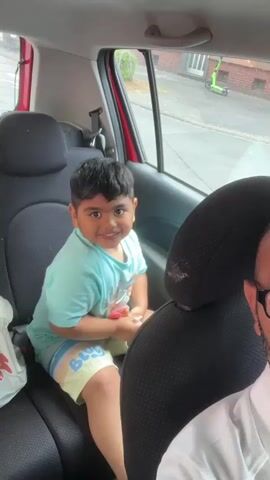 A young boy with dark hair and a light blue t-shirt and yellow shorts sits in the back seat of a red car. He is smiling and holding a red and yellow object, possibly candy. To the right of the boy, the headrest of the front passenger seat is visible, and a person wearing glasses and a white shirt is partially in view. Outside the car window, a street scene in Dortmund, Germany, is visible, including buildings, a sidewalk, and a green electric scooter. The lighting suggests it is daytime.
