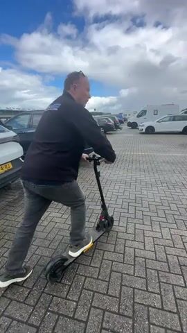 A man rides an electric scooter through a paved parking lot. He is wearing a black jacket and gray pants. The scooter is black with white accents. Several cars are parked in the lot, including a dark gray SUV to the left and a white car to the right. The sky is cloudy with patches of blue.