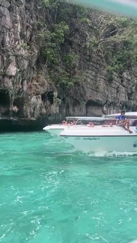 A motorboat with several passengers moves through clear turquoise water near a rocky, jungle-covered cliff face. The boat is white with blue accents and displays "PORNPHINYO MARINE" and the number "959" on its side. Two Thai flags are visible on the boat's canopy. One person sits at the front of the boat, while others are seated or lying down inside. The water is choppy and reflects the bright sunlight.