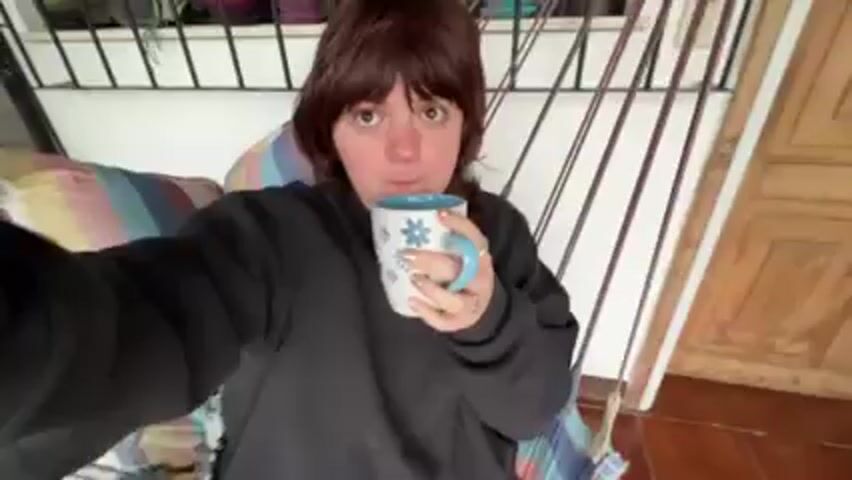A young woman with short, dark hair, wearing a dark sweatshirt, is holding a white mug with blue flowers and drinking from it. The camera is close to her face, suggesting she is taking a selfie. The background includes a cushioned seat, a white railing with plants, and a partial view of a door. The scene then pans upwards to show a view of a lake or ocean with mountains in the distance, seen through an arched opening. There are potted plants along the edge of the patio and a decorative, dark sculpture on the wall. The floor is made of reddish-brown tiles. The video is slightly shaky as if hand-held.