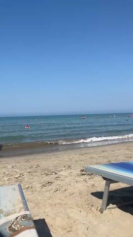 The video shows a sunny day at the beach. The sand is visible in the foreground with two lounge chairs, one in focus on the left and another partially visible on the right. Beyond the sand, the ocean stretches to the horizon under a clear blue sky. Gentle waves are breaking on the shore. Several people are in the water, some swimming and others appearing to be floating. One person, a child wearing a hat and shorts, runs into the water from the right side of the frame. The overall scene depicts a peaceful and recreational beach environment.
