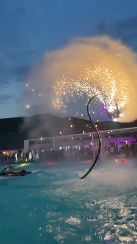 The video shows a nighttime water show. A person is suspended in the air above a pool of water, propelled by a jet ski and a water-powered jetpack. Sparks are erupting from the jetpack, creating a dazzling display against the twilight sky. A crowd of people watches from the poolside, some holding up phones to record the event. The background features hills and a building illuminated with lights. The water is a bright blue, reflecting the lights and sparks.