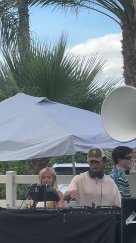 The scene depicts a daytime outdoor party setting, likely a pool party or similar event. Focus is on a DJ booth where a person in a beige outfit and a baseball cap is actively DJing, using Pioneer DJ equipment. A woman with blonde hair stands beside the DJ, holding a drink, seemingly enjoying the music. Several people are standing around, some holding drinks and engaging in conversation, giving the impression of a social gathering. The environment includes palm trees providing shade, a large white umbrella for shade as well, and a large white horn loudspeaker visible. The weather seems pleasant, with a blue sky and scattered clouds. Overall, the video shows a lively outdoor event centered around music and social interaction.