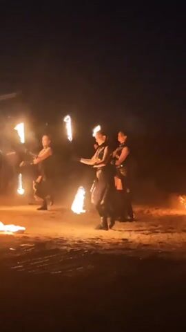 Three women are performing a fire dancing routine in an outdoor, dimly lit area. They are dressed in dark, revealing outfits with boots and appear to be holding and manipulating torches or poi that are on fire. The ground is dirt and there are several small fires burning on the ground around them. The background is dark, suggesting it is nighttime or dusk. One woman is in the foreground on the left, another is in the middle, and the third is on the right. The woman in the middle is actively spinning fire props. There is some smoke visible in the air from the fires.