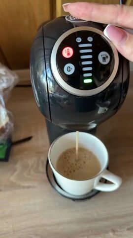 A hand with manicured nails presses a button on a black coffee maker. A thin stream of coffee is pouring into a white mug with a silver rim, which sits on a matching saucer. The coffee maker has a circular control panel with various illuminated icons and indicators. A red thermometer icon is illuminated, as is a green light at the bottom of a vertical series of bars. The coffee maker sits on a wooden countertop.