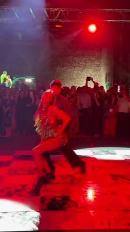 This video shows two dancers, a man and a woman, performing on a checkered dance floor under red stage lighting. The woman wears a short, sparkly green dress, red tights, and black boots, and her hair is dark and appears to be styled with volume. The man is dressed in a teal collared shirt and black pants. The background shows an audience watching the performance, with some individuals holding up their phones. The environment appears to be an indoor event space with brick walls and a ceiling with visible lighting rigs. The dancers are in the foreground, engaged in synchronized movements, with the woman often leading with dynamic gestures.
