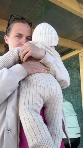 A woman holds a baby in her arms in an outdoor setting with a wooden structure overhead. The woman is wearing a light-colored jacket, a pink top underneath, and sunglasses on her head. Her hair is pulled back. She's holding the baby, who is dressed in a light-colored knit outfit and hat. She lifts the baby into the air, smiles, and then brings the baby back down into her arms. To the side, there is a light green baby stroller. The structure above has wooden beams and a green-patterned covering. The background appears to be a building or shed with a dark door.