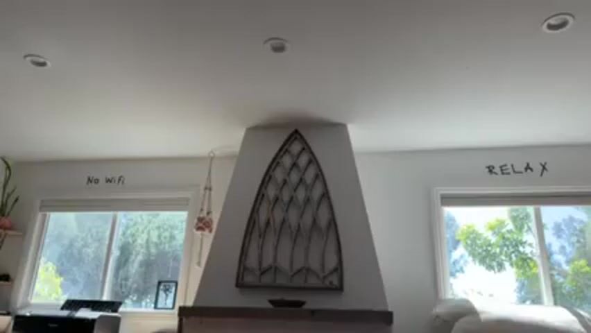 The video shows a room with white walls, two windows, and a fireplace. The wall above the left window has the words "No WiFi" written on it. A hanging plant is in front of the window. The wall above the right window has the word "RELAX" written on it. The fireplace has a decorative gothic-style wooden piece on the wall above it.