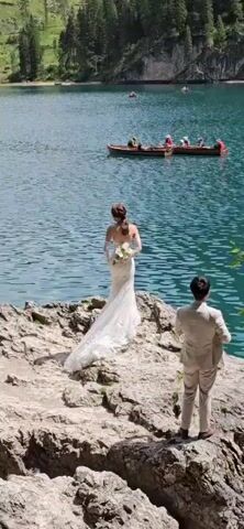 A bride in a white wedding dress stands on a rocky shore, facing a body of blue water. She holds a white bouquet. Behind her, a man in a light-colored suit stands with his back to the camera, possibly taking a photo. In the background, two wooden boats are on the water, with several people in them. The water is calm and reflects the sky. The sun is shining, casting shadows on the rocks.