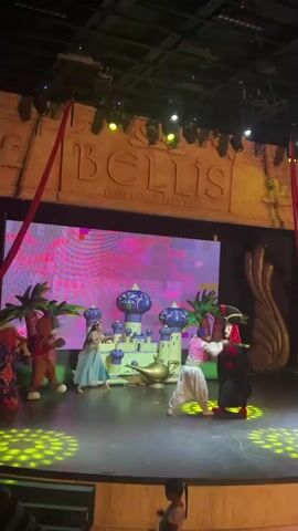 A stage performance is underway, likely a re-enactment of Aladdin, given the visual cues of a castle with domes, palm trees, and a magic lamp. The backdrop features a large screen displaying abstract, colorful patterns and the hotel's name, "BELLIS Deluxe Hotel." Red lighting illuminates the stage, with spotlights highlighting the performers. In the foreground, three costumed characters are on stage. To the left, a character in a purple and gold outfit with a headdress is partially visible. Center stage, a female character in a flowing blue costume, presumably Princess Jasmine, interacts with two other characters. One is a male character in a white outfit, appearing to be Aladdin. The other is a villainous character dressed in black and red with a distinct headpiece, likely Jafar. Jafar appears to be holding Aladdin. Red fabric ribbons hang from the ceiling, adding to the theatrical ambiance. In the lower portion of the frame, a young girl is visible from the back, seemingly watching the show. The stage itself is dark wood, with steps leading up to it.
