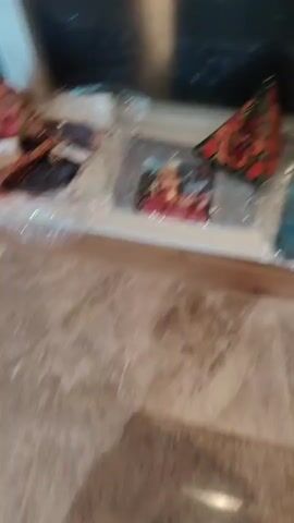 The video pans across a display of various bags and accessories. The items appear to be in clear plastic packaging, suggesting they are new or being sold. The display is set up on what looks like a shelf or counter, with a marble-patterned surface below. The lighting is somewhat dim, creating reflections on the plastic packaging. Some of the items are colorful, featuring floral patterns and embroidered designs. One item in the foreground is a small, triangular-shaped pouch with a floral print and a keychain attached. There is also a small rectangular box with a tag. Further back, more bags are visible, including some with bold embroidered designs and others with subtler patterns. The overall impression is of a retail display of handmade or artisanal bags and accessories.