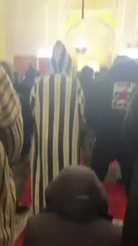 The video shows a group of people inside a mosque, apparently praying. The viewpoint is from the back of the crowd, focusing on several people. One person is wearing a striped hooded djellaba, another a black hoodie with "JORDAN 23" on the back. The interior architecture of the mosque is visible, featuring arched doorways and intricate wall patterns. The floor is covered with red carpeting. The audio suggests a religious chant or prayer.