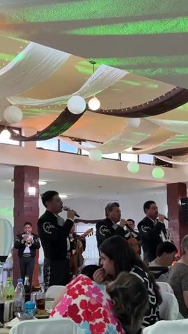 A group of four men dressed in traditional Mexican mariachi outfits are performing at a gathering. Three of the men are singing into microphones while playing acoustic guitars, and the fourth man is playing a trumpet. The audience is seated at tables, watching the performance. One woman in a colorful floral shirt sits near the front, and a young child sits on someone's lap nearby. The room is decorated with white fabric draping from the ceiling, accented with green lights, brown streamers, and white paper lanterns. There are also strings of decorative lights. The room has a festive and celebratory atmosphere.