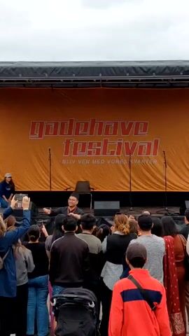 The video captures a performance at the Gadehave Festival. A crowd of people is gathered in front of a stage, many holding up their phones to record. On the stage, a man is seated, wearing glasses and a hat. A woman in a baseball cap is kneeling to his left. A large orange banner with the festival's name is draped behind them. Speakers flank the stage.