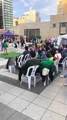 A large crowd of people is sitting in white plastic chairs in an outdoor setting, facing a stage area with tents. The building "UMSA" is visible in the background. Many people are looking towards the stage, some raising their arms and clapping. Several individuals hold up a Bolivian flag. The weather is overcast, and the ground is paved with square tiles. Some people are standing at the edge of the seating area.