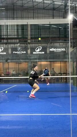 A group of people are playing padel tennis in an indoor court. The court is blue with white lines. There is a net in the middle of the court. In the foreground, a woman with dark hair tied in a bun is wearing a black t-shirt with "MUSCLE" printed on the back in white letters, and black shorts. She is holding a padel racket and is in the process of hitting a yellow ball. She is wearing pink shoes and white socks. In the background, on the other side of the net, another woman is also playing padel tennis. She is wearing a dark t-shirt and red shorts. Two other people, a man and a woman, are standing behind the net on the left side of the court. The court is surrounded by glass walls and a metal structure. Banners for "Padel Club", "DECATHLON", and "Niko Padel Academy" are visible on the wall. The lighting is artificial.