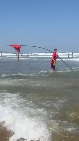 A person in a red shirt and red shorts stands in shallow ocean water holding a long black pole with a red flag attached to the end. The flag is unfurled and flapping in the wind. The person is also holding a yellow flotation device or buoy. In the background, several other people are in the water, some swimming and some standing. The sky is blue and clear, and there are waves breaking on the shore. The scene appears to be a beach on a sunny day.