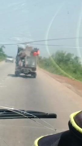 The media file shows a view from inside a vehicle, looking out through the windshield. The windshield has some scuffs and streaks on it. In the distance, a truck is driving away on a paved road. The truck appears to be overloaded with what looks like cargo and possibly people. The truck has a dark color and its brake lights are illuminated. The road is bordered by dry grass and trees, suggesting a rural or semi-rural setting. The sky is bright and clear, indicating it is daytime. The lighting suggests it might be late morning or early afternoon. The overall impression is a snapshot of travel on a road in Nigeria.