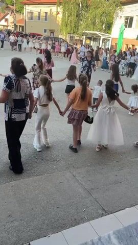 A group of adults and children hold hands and dance in a circle on a paved area. Many other people are standing around in the background, some of whom are also dancing in a larger circle. The scene appears to be an outdoor event, possibly a celebration or festival, with buildings visible in the background.