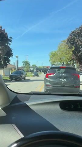 The view from inside a car shows traffic stopped at a railroad crossing. The train consists of gray tank cars. A black Honda SUV is stopped on the left side of the road in front of the car. A GMC SUV is directly in front of the camera. The sky is blue and sunny. The railroad crossing signals are activated.