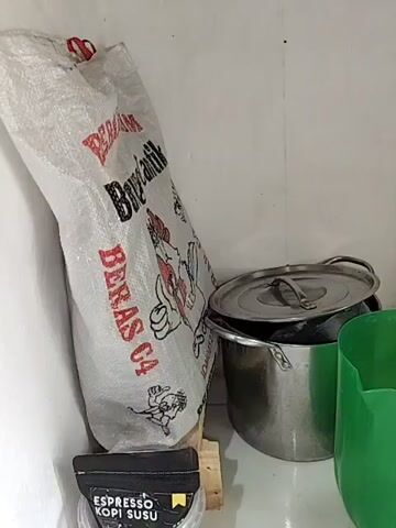 The video shows a corner of a room, possibly a kitchen or storage area. In the foreground, to the left, is a large white sack with red lettering that appears to be rice. Below the sack is a black package with white lettering that says "ESPRESSO KOPI SUSU". To the right of the rice sack is a metal pot with a lid, and next to that is a bright green plastic bucket. Further to the right, a white bucket with a red and blue label is visible. In the background, a white wall with some pipes and a sink drain are present. The lighting is dim.