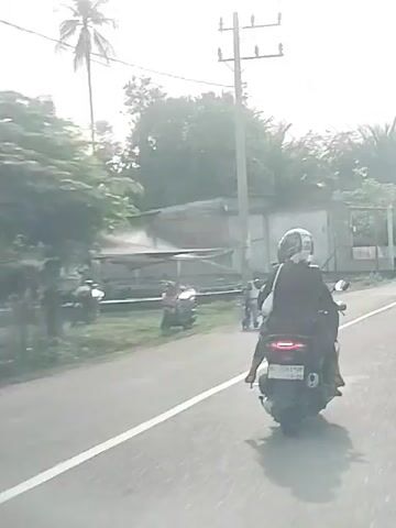 The video shows a rear view of a motorcycle with two people on it, riding on a paved road. The person in the back is wearing a dark colored outfit and a helmet. In the background, on the left side of the road, a person in a wheelchair is visible. The environment appears to be tropical with palm trees and other vegetation visible. The sky is bright, suggesting daylight.