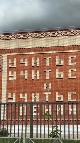 The camera is positioned at a low angle, slightly behind a grey metal fence, providing a clear view of a brick building with a notable message embedded into its exterior. The facade is primarily constructed of orange-red bricks arranged in a grid-like pattern, with a horizontal border of tan bricks across the top. A large rectangular space in the center of the brickwork features white bricks arranged to spell out a Russian phrase across multiple rows, partially obscured by the fence. A small green plant grows near the base of the fence, adding a touch of nature to the scene. Overhanging the scene, a dark grey, cloud-filled sky conveys an overcast day.