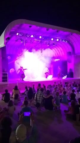 This is a wide-angle, low-resolution video recording of an outdoor performance at night. A stage with a white canopy is lit by purple and blue lights and filled with smoke. Three performers, two women and one man, are on stage, dressed in theatrical costumes. The women are wearing dark dresses, and the man is wearing a dark suit. They are holding umbrellas, and there are large, white, spherical objects on the stage. A crowd of spectators, including children, are seated on the ground in front of the stage, watching the performance. Some audience members are holding up phones to record the event. The atmosphere is festive and celebratory.