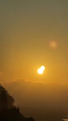 This media file captures a scenic journey through Gülnar, Turkey. The video begins with a breathtaking view of a sunset over a mountainous landscape, with the sun appearing as a bright, double-orb due to a lens flare. Lush trees dot the hills, and a body of water is visible in the distance. The scene then transitions to the interior of a moving vehicle, likely a bus, as indicated by the visible seats and windows.

The camera pans across the passing scenery, showcasing a winding road and glimpses of arid hillsides dotted with vegetation. A highway tunnel entrance appears in the distance, marked by a directional sign with a red arrow. The time of day is clearly either sunrise or sunset, as indicated by the warm, golden hues illuminating the sky and landscape. The weather appears clear and pleasant, ideal for travel. While no people are explicitly shown, the perspective from within the vehicle suggests passengers are present, enjoying the journey. The focus is on the visual experience of the landscape and the transition through different geographical features, creating a sense of movement and exploration. There is no visible text in the media file.