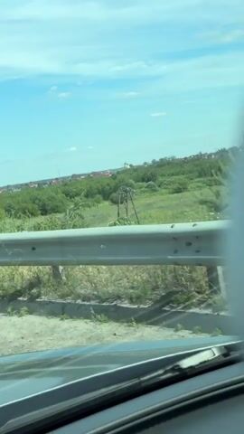 This video appears to be a first-person perspective from inside a vehicle traveling along a road. The primary visual focus is the guardrail that runs alongside the road. Beyond the guardrail, there is a landscape of green fields and trees, with some buildings and a steeple visible in the distance, suggesting a rural or semi-rural setting. The sky is blue with scattered white clouds, indicating fair weather and likely daytime, possibly late morning or early afternoon given the brightness.

The activity captured is the movement of the vehicle, indicated by the changing scenery and the perspective from within the car, including a view of the dashboard and windshield wipers. There are no people visible in the frame. The guardrail is a prominent object, constructed of metal poles and horizontal bars, and it runs along the edge of the road, separating it from the fields.

The location is identified as Orlivka, Ukraine. There are no discernible signs or text visible in the video that would provide additional information. The overall mood conveyed is one of travel and observation of the passing landscape.