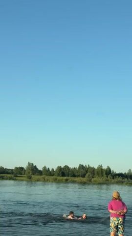 The video shows a wide, calm river under a clear blue sky. On the left side of the river, two people are swimming. On the right bank, a person wearing a pink shirt and patterned shorts stands with their back to the camera, looking out at the river. Trees and greenery line the far bank of the river, creating a natural landscape. The scene appears to be a peaceful outdoor setting.
