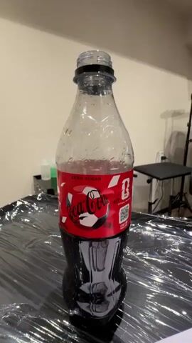 A close-up video shows a bottle of Coca-Cola Zero Sugar with a soccer-themed label. The bottle is partially filled with dark liquid and condensation is visible on the clear plastic. The label features the Coca-Cola logo, the words "ZERO SUGAR," and a soccer ball design. A QR code is also present on the label. The bottle is placed on a dark, reflective surface that appears to be covered in plastic sheeting. In the background, there are shelves with bottles and equipment, suggesting an indoor setting, possibly a studio or workspace. The lighting is consistent, and the camera remains focused on the bottle as it slowly zooms out.