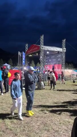 The video shows an outdoor evening event with a stage set up in the background. A large banner behind the stage reads "STAYS 2026" and appears to be advertising an event. There are metal truss structures supporting the stage and lighting equipment. Several tents are visible in the background. A crowd of people are gathered in the area, some standing and some sitting. A young boy in a blue jacket and white pants walks past the camera from left to right. To his right, an adult in a dark tracksuit and a cap walks in the same direction, arms crossed. Other individuals are visible in the middle and background, some appearing to be participants or spectators. The ground is covered in grass and dirt, and the sky is dark, suggesting it is nighttime. The overall scene appears to be a sports or entertainment event held outdoors in the evening.