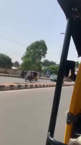 The video shows a view from inside a yellow auto rickshaw moving along a paved road on a bright, sunny day. The road has a dividing median with alternating black and white curbs. In the background, there are several palm trees and other greenery. There are also buildings, some painted white and some darker. Other vehicles, including a motorcycle, are also visible on the road.  A person is seen standing on the side of the road. The camera is moving forward, as the auto rickshaw is in motion.