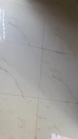 The video shows the interior of a room undergoing renovation. The floor is tiled with large white tiles with marble-like patterns. In one view, a pair of yellow rubber boots are on the floor, along with a bag of construction material and a metal mesh. The camera then pans to an adjacent shower area with tiled walls and floor, also featuring a marble-like pattern in grey tones. The walls of the shower area are partially finished, with some exposed areas showing the underlying wall structure. There are no people visible in the video.