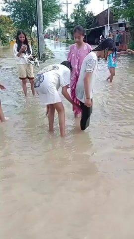 The video shows a flooded street in a residential area. Five young women are wading through the water, and a young boy in a black shirt and red shorts is running in the water behind them. Other people, including a man in a red shirt and a younger child, are also seen in the water. The water is murky and brown, and it appears to be several inches deep. Houses and trees line the street, and utility poles are visible in the background. The women are wearing casual clothes, and some of them are smiling and laughing. The overall tone of the video is lighthearted, despite the flooding.