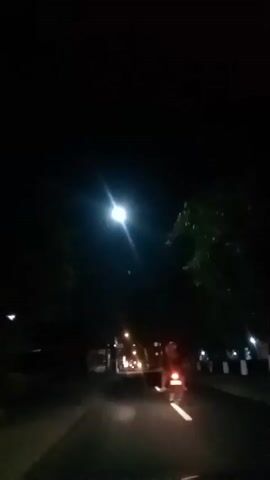 A nighttime video captures a street scene from a moving vehicle. The camera is looking forward, down a paved road lined with trees and buildings. Streetlights illuminate the road, casting a warm glow. A motorcycle is visible ahead, its taillight glowing red. The sky is dark, with a bright light, possibly a streetlight or the moon, visible in the upper portion of the frame. The overall impression is one of a quiet, urban night.