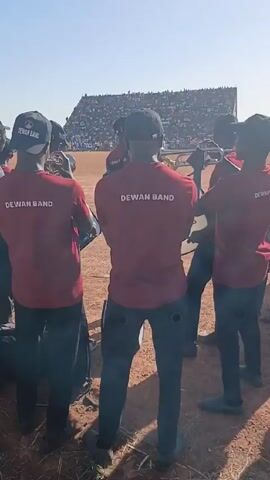 A group of musicians from the "DEWAN BAND" are standing on a dry field in front of a stadium filled with spectators. The musicians are wearing matching red t-shirts with "DEWAN BAND" printed on the back and black pants. Some are playing trumpets, and one is holding drumsticks near a bass drum. A young boy in shorts and sandals stands slightly apart from the group. The sky is clear and the sun is shining.
