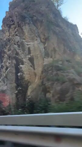 The video begins with a blurry shot of a road sign indicating a sharp left turn, with a rocky cliff face in the background and a clear blue sky above. The camera then pans to show a mountainous landscape with lush green trees and a winding road. The perspective shifts to the interior of a car, showing the back of a person's head and shoulders as they sit in the passenger seat. The camera then focuses on the driver, who is holding up a smartphone to record the road ahead. The driver is wearing glasses and a black t-shirt, and the car's dashboard and steering wheel are visible. The smartphone screen shows a navigation app. The car is moving along a scenic road with steep, rocky cliffs on one side and a dense forest on the other. The driver's hand is visible on the steering wheel. The video ends with a shot of the driver's head and shoulder from behind as they continue to drive.