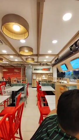 The video pans across the interior of a brightly lit restaurant, likely a casual dining establishment, possibly a fast-food chain given the prominent digital menu boards. The decor features a modern aesthetic with light wood accents and white ceilings with exposed wooden beams.

Several individuals are visible. In the foreground, a person's head and shoulders are seen from behind, looking down. Further into the restaurant, a young boy in a striped shirt is seated at a table, also looking down, possibly at a device. In the background, several restaurant staff members in uniform are visible behind a counter, interacting with customers or preparing food. Other diners can be seen at tables in the middle ground.

The scene appears to be during daylight hours, as indicated by the overall brightness of the interior. The setting is clearly a food establishment, with tables and chairs arranged for dining. The activity taking place is typical of a restaurant: people dining, staff serving, and a general hum of activity associated with a meal service.

Notable details include the digital menu screens displaying various food items, one prominently featuring a bowl of beef. The restaurant name "DIMSUM DRINK" is visible in large letters on a sign in the middle ground. The overall atmosphere seems relaxed and functional, typical of a quick-service restaurant. The location cues such as "Cebu City, Philippines" provide geographical context.