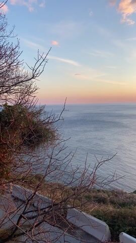The video captures a breathtaking sunset view over a vast body of water, likely an ocean, from a high vantage point on a cliff or hill. The scene begins with a wide shot of the calm, blue-grey water extending to the horizon, meeting a sky painted in soft gradients of blue, pink, and orange. Bare, dark tree branches with fine twigs are prominent in the foreground on the left side, indicating a foreground element.

As the camera slowly pans to the right, more of the coastline comes into view. The land below is rugged and rocky, covered with patches of dry grass and low-lying vegetation. A small cluster of dark rocks forms a tiny islet or outcrop closer to the shore.

The pan continues until the vibrant, orange sun becomes visible, sitting low on the horizon. It casts a brilliant, shimmering path of light across the tranquil water, creating a strong sunbeam effect. The sky around the sun is intensely orange and yellow, fading into softer pinks and blues higher up. In the final frames, additional dry grass and a white barrier or railing are visible in the immediate foreground on the right, framing the dramatic sunset. The overall atmosphere is peaceful and majestic, showcasing the natural beauty of the sunset over the sea.