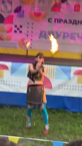 A young woman with dark hair tied back and wearing a brown vest, a brown skirt with fringe, blue leggings, and red shoes is performing with fire. She is on a grassy area in front of a stage with a banner that says "с праздником ДВУРЕЧЬЕ!". She is spinning what appear to be poi, which are flaming balls attached to ropes, around herself. Her body is angled slightly to the left, and her arms are extended. The background is blurred and shows stage lighting and a colorful banner. In the foreground, the heads and shoulders of an audience are visible.