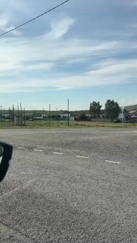 The video shows a view from a vehicle traveling on a paved road. The road has white lane markings. On the right side of the road, there are various structures including utility poles, small buildings, and some fencing. The background consists of a grassy field with scattered trees and houses. The sky is blue with scattered clouds. The video appears to be taken during the daytime.