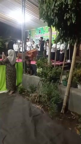 The video shows an outdoor event at night. A group of young men in white shirts and pants are standing on a stage decorated with green banners. One man in the center appears to be speaking into a microphone. To the left, a woman wearing a hijab and a patterned dress is standing and holding up a phone, possibly recording the event. In the foreground, there are plants and trees. The overall scene suggests a community gathering or performance.