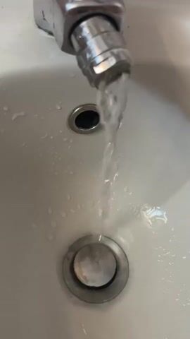 This is a close-up video of a faucet running water into a white sink. The water flows from a silver faucet with a textured grip and streams down into the sink basin, passing over a smaller drain opening before reaching a larger drain with a chrome stopper. The sink surface is wet with droplets of water scattered around. The lighting suggests it might be daytime, but there are no explicit clues about the weather. The setting appears to be a bathroom or kitchen sink. The activity is simply water running from a faucet. There is no visible text in the video. The location is noted as Varese, Italy, but this context is not visually evident within the frame itself.
